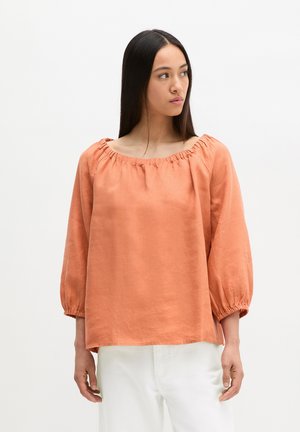 Femme aux longs cheveux noirs portant un chemisier orange à épaules dénudées et un pantalon blanc, regardant vers sa droite sur un fond uni.