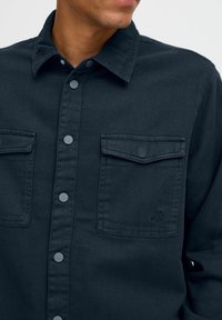 Dark denim shirt with buttoned front, two chest pockets, and a collar. Features a subtle logo on the left pocket and textured fabric.