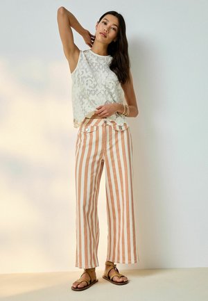 Nő ujjatlan fehér csipkefelsőben, magasderekú barack és fehér függőleges csíkos nadrágban és bézs szandálban, egy emelt karral áll.