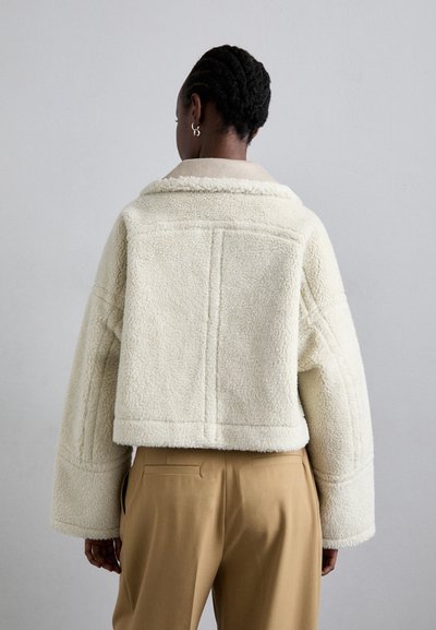 Geknipte schapenvachtjack in crème, met een brede kraag, verlaagde schouders en textuurstof. Gecombineerd met beige op maat gemaakte broeken.