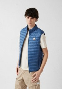 Chaleco acolchado azul, diseño acolchado, cuello alto, cremallera frontal, parche con logo en el pecho. Llevado sobre una camiseta beige, combinado con pantalones cargo kaki.