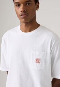 White cotton t-shirt with short sleeves, featuring a left chest pocket and a red graphic label. Smooth texture, relaxed fit.