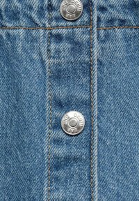 Světle modrý denimový materiál s viditelnou strukturou. Obsahuje stříbrné kovové knoflíky a kontrastní žluté stehy podél švů.