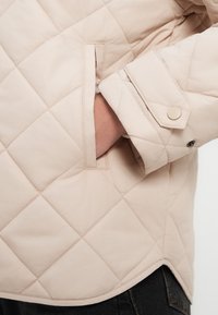 Μπεζ καπιτονέ μπουφάν με διαγώνιο σχέδιο, διαθέτοντας πλαϊνή τσέπη και κούμπωμα με κουμπιά στο μανίκι. Λείο υλικό και μοντέρνος σχεδιασμός.