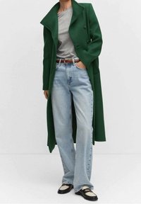 Long manteau vert à revers crantés et boutons croisés, associé à un t-shirt gris, un jean bleu clair et des chaussures noires et blanches.