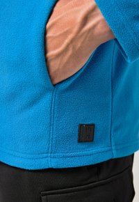 Niebieska kurtka z polaru o fakturowanej powierzchni, z boczną kieszenią i czarną naszywką z logo przy dolnym brzegu. Poniżej widoczne czarne spodnie.