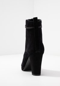 Botín de ante negro con tacón alto, que presenta un puño doblado y una correa decorativa. Textura suave y silueta distintiva.