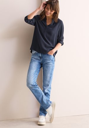 Marineblaue Bluse mit langen Ärmeln und Knopfleiste, kombiniert mit hellblauen geraden Jeans und weißen Sneakers.