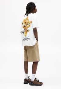 Weißes Baumwoll-T-Shirt mit einer Grafik eines gelben Blumenvasens auf dem Rücken, kombiniert mit beigen Shorts und braunen Sneakers mit schwarzen Akzenten.