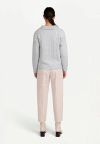 Ljusgrå stickad tröja med kabelstickat mönster, ribbad nederkant och krage, kombinerad med beige byxor. Modellen ses bakifrån.