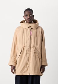 Beige vanntett parka med stor krage, snøring i midjen og to frontlommer. Glatt tekstur; har et rosa merke festet.