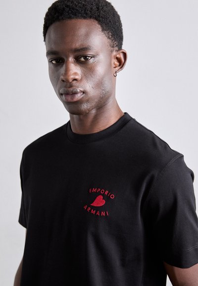 T-shirt en coton noir avec un logo brodé rouge sur la partie gauche de la poitrine, col rond et manches courtes. Texture de tissu lisse.