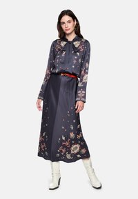 Marineblaues Kleid mit Blumenmuster, langen Ärmeln und einer Schleife am Hals. Verfügt über einen unifarbenen Taillenbund. Kombiniert mit weißen Stiefeln mit Absatz.