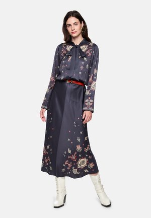 Marineblaues Kleid mit Blumenmuster, langen Ärmeln und einer Schleife am Hals. Verfügt über einen unifarbenen Taillenbund. Kombiniert mit weißen Stiefeln mit Absatz.