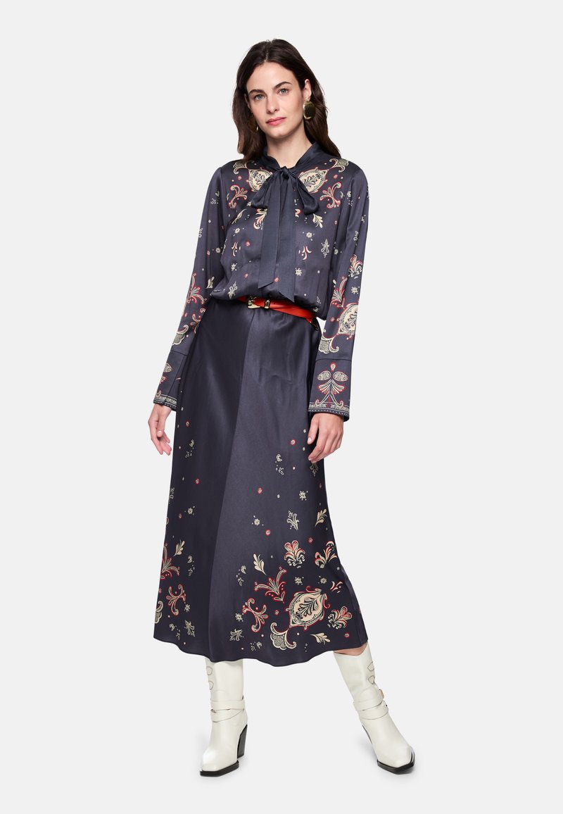 Marineblaues Kleid mit Blumenmuster, langen Ärmeln und einer Schleife am Hals. Verfügt über einen unifarbenen Taillenbund. Kombiniert mit weißen Stiefeln mit Absatz.