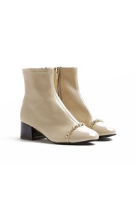 Béžové kotníkové boty s lesklou lakovanou špičkou, řetízkovým akcentem a středně vysokým blokovým podpatkem. Mají centrální zip a hladkou texturu.
