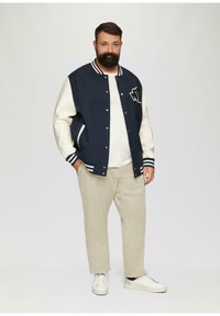 Marineblauw en crèmekleurig varsityjack met een letterpatch, gestreepte boorden en zijzakken, gecombineerd met beige broek en witte sneakers.