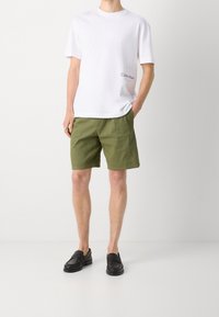 Biała bawełniana koszulka z małym czarnym logo, w połączeniu z zielonymi szortami i czarnymi mokasynami z teksturowanym wykończeniem oraz zakrytymi palcami.