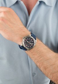 El reloj de pulsera para hombre presenta una caja de acero inoxidable, correa de goma azul, esfera negra con números blancos y rosas, y subesferas de cronógrafo.