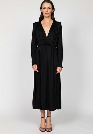 Kobieta stoi, ubrana w czarną sukienkę z długim rękawem, głębokim dekoltem w kształcie litery V, taliowaną, oraz czarne szpilki z paskiem wokół kostki, na jednolitym tle.