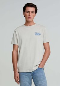 Light beige cotton t-shirt with short sleeves. Features a blue embroidered "Scalpers" logo on the left chest. Classic crew neck design.