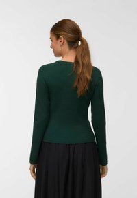 Haut long à manches ribbées vert foncé avec une silhouette ajustée et un col rond, associé à une jupe noire fluide. Tissu texturé visible.