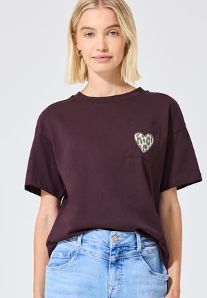 Jeune femme portant un t-shirt violet foncé avec un cœur imprimé léopard sur la poche poitrine et un jean bleu clair.