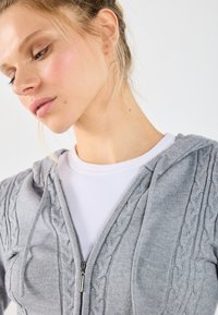 Gray knitted zip-up hoodie with a cable pattern, featuring a hood and zip closure. Worn over a white ribbed shirt. Smooth texture.