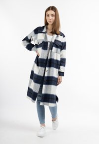 Cappotto a quadri blu e bianco con bottoni, dotato di due tasche sul petto, maniche lunghe e una vestibilità comoda, abbinato a jeans azzurri e sneaker bianche.
