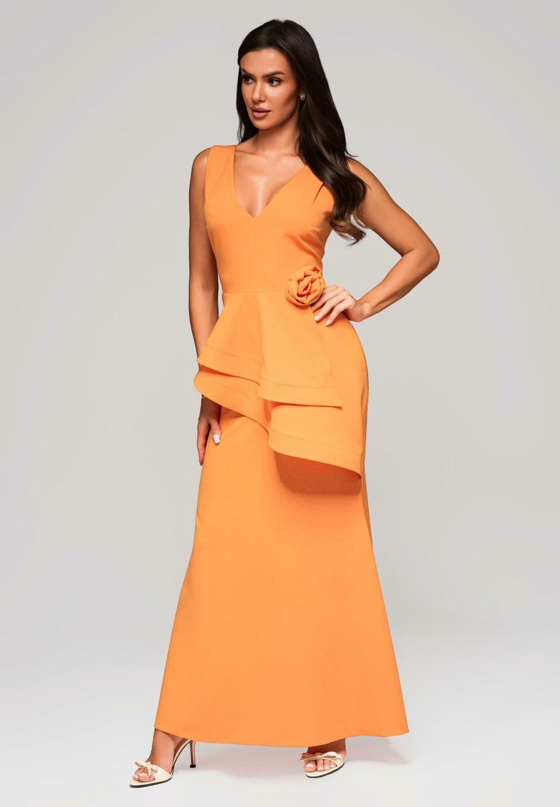 Vrouw met lang, donker haar in een mouwloze oranje jurk met gelaagde ruches en een bloemdetail op de taille, poserend met haar hand op de heup.