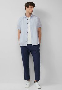 Helles blaues kurzärmeliges Hemd mit Knöpfen, weißes T-Shirt darunter, dunkelmarineblaue schmal geschnittene Hose und weiße Sneakers mit schwarzen Akzenten.