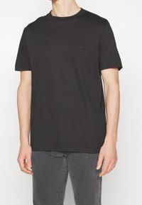 Czarny t-shirt z krótkim rękawem i okrągłym dekoltem, z małym kwadratowym logo na lewej piersi, noszony z wyblakłymi czarnymi dżinsami, widoczna sylwetka od podbródka do bioder.