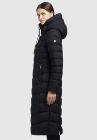 Palton puffer negru cu un design lung, ajustat, glugă mare, buzunare laterale cu fermoar și textură matlasată cu detalii de cusătură contrastante.