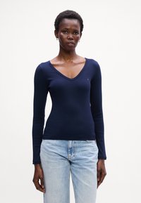 Tricou cu mânecă lungă și decolteu în V de culoare bleumarin, fabricat dintr-un material ribbed. Are o siluetă ajustată și o mică aplicație logo pe pieptul stâng. Este asociat cu blugi de culoare albastru deschis.