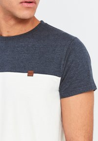 Mænds T-shirt med korte ærmer i mørkeblå og hvid, der har en struktureret stof, rund halsudskæring og et lille brunt mærke på brystet.