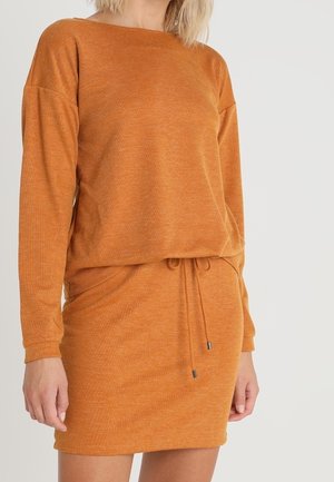 Žena v dlouhých rukávech, oranžových pletených šatech s šňůrkou v pase, zobrazená od krku po polovinu stehenní oblasti na jednobarevném pozadí.