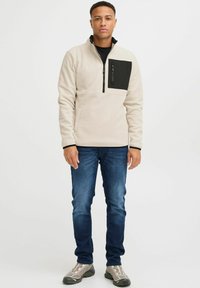 Sudadera de forro polar beige con un bolsillo en el pecho de cremallera negra de un cuarto; cuello y puños negros; combinada con jeans azules y zapatos grises.