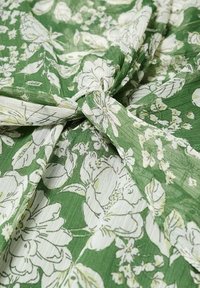 Tissu vert avec imprimé floral blanc, rassemblé et noué au centre, montrant la texture et les détails fins des fleurs.