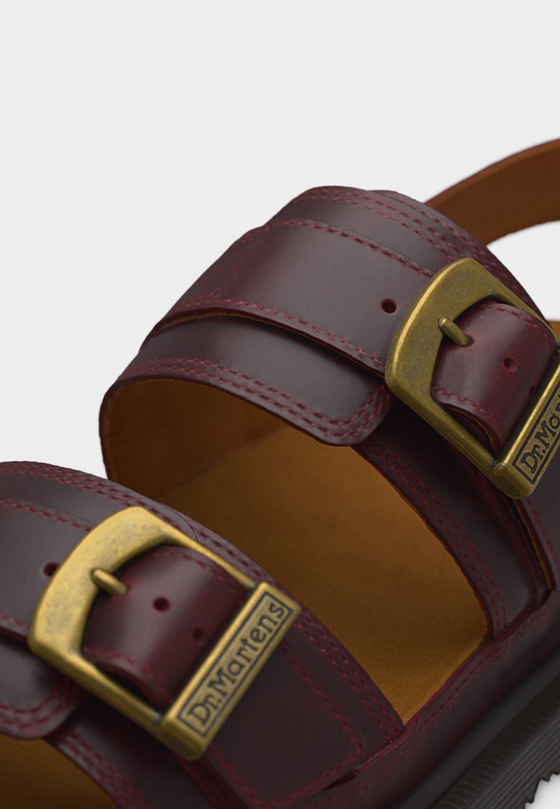 Nahaufnahme von dunkelroten Leder-Sandalenriemen mit Messingschnallen, die mit "Dr. Martens" beschriftet sind, und detaillierter Nahtarbeit.