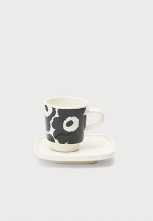 Tasse en céramique blanche avec un motif floral noir sur une soucoupe carrée blanche assortie, sur un fond clair uni.