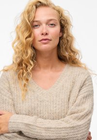 Bež pulover z V-izrezom, rebraste teksture in ohlapnega kroja. Pletivo je mehko in puhasto na videz, z dolgimi širokimi rokavi.