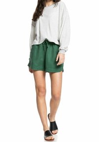 Langärmliges Oberteil in hellgrau und weißer Streifenoptik, kombiniert mit grünen Shorts mit Kordelzug. Das Model trägt schwarze Rutsch-Sandalen.