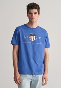 Tricou din bumbac albastru, cu guler rotund, mâneci scurte și un design grafic cu un logo în formă de scut și textul "1949 GANT AMERICAN SPORTSWEAR".