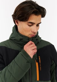 Giacca puffer verde e nera con colletto alto, tasca con zip arancione e polsini in Velcro. Presenta una cappuccio regolabile e tessuto testurizzato.