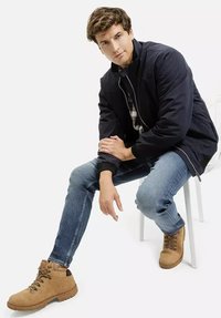 Granatowa kurtka na wierzchu koszuli w kratę, niebieskie dżinsy i tanie sznurowane buty. Model siedzi na białym krześle, prezentując wyluzowaną postawę i swobodny styl.