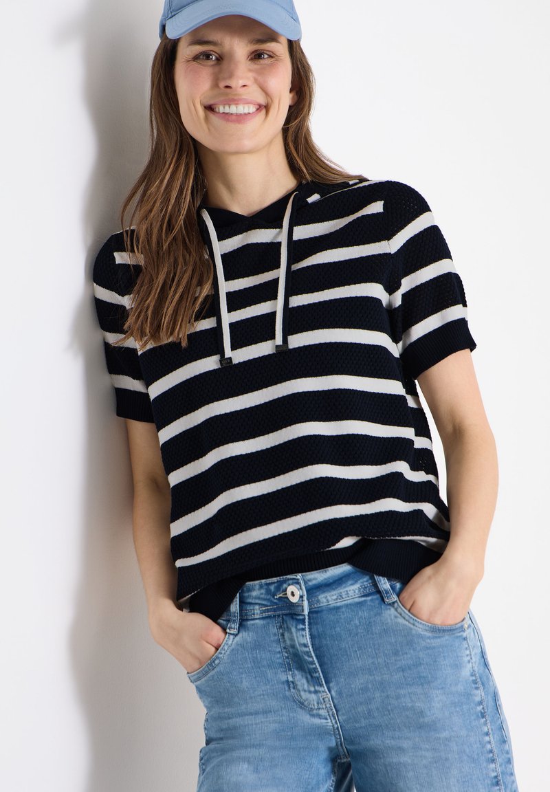 Femme souriante appuyée contre un mur, portant un sweat à capuche à manches courtes rayé noir et blanc, une casquette de baseball bleue et un jean bleu clair, les mains dans les poches.