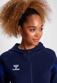 Mörkblå zip-up hoodie med en logotyp, som har kontrasterande blå detaljer, mjukt tyg och en rundad krage.