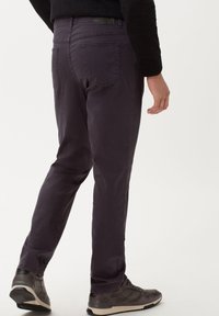 Uomo che indossa pantaloni dritti viola scuro con tasche laterali e posteriori, abbinati a scarpe da ginnastica nere e a un top nero a maniche lunghe.