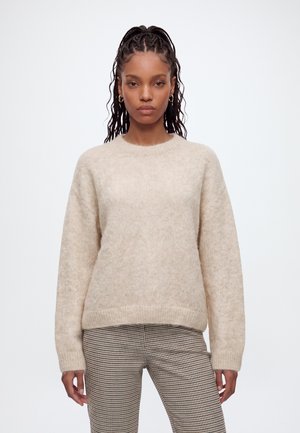 Ung kvinde med flettet hår iført guldringeøreringe, en beige sweater og sort-hvide ternede bukser, stående mod en hvid baggrund.
