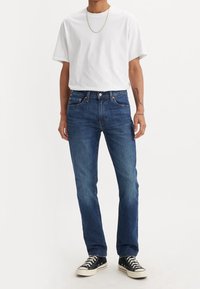 Personne portant un t-shirt blanc uni, un jean bleu droit, des baskets noires avec des lacets blancs et une fine chaîne en or.